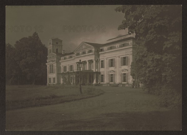 Werki. Palace - facade, Bulhak, Jan 1876–1950 photographer, Vilnius Lithuania, architecture, residential architecture, palaces, ca. 1920, photography, nature photography, height 11.5 cm, width 16.0 cm, A large classical building with columns and a triangular pediment sits slightly right of center, flanked by a taller tower-like section on the left and dense trees on the right and left; a lamppost stands on a grassy area in front of the building beside a curved dirt path, and the scene is rendered in muted tones of brown, sepia, green, and dark gray.