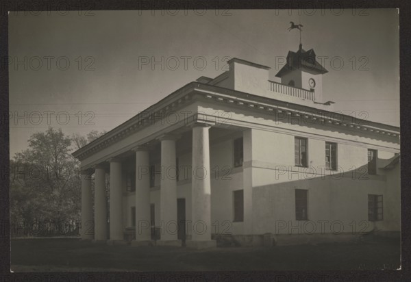 Stable, architecture, stables; photographer: Jan Bulhak 1876–1950; subject: Werki, Vilnius Vilnius, Lithuania; ca. 1934; photograph; nature photograph; dimensions: height 11.2 cm, width 16.7 cm., A large classical building with a columned portico occupies the foreground, showing a pale exterior with tall round columns supporting an entablature and a roofline with decorative dentils; the façade has several rectangular windows and door openings, and a smaller upper structure with a cupola topped by a weather vane rises above the roof. The scene includes a group of trees to the left, an open sky above, and areas of light and shadow playing across the building surfaces; visible colors include shades of gray, off-white, black, and muted dark green in the trees.
