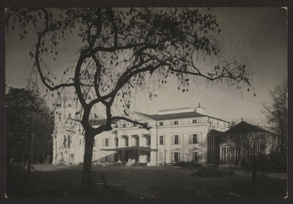 Werki — palace facade; photographer: Jan Bulhak 1876–1950; subject: palaces in Werki Lithuania, Tyszkiewicz family residence; noted by Mieczyslaw Orlowicz 1881–1959; circa 1934; location: Vilnius; nature photography; dimensions: 11.3 × 16.9 cm., A large dark tree with many thin hanging branches and sparse leaves dominates the foreground, its silhouette contrasting against a lighter sky; a simple wooden bench sits near the tree trunk. Behind the tree, a grand multi-story building with a classical facade and columned portico extends across the scene, flanked by a glass-roofed conservatory on one side. The ground in front of the building is a grassy lawn with scattered low plants and shadows. Visible colors include black, dark gray, light gray, and muted off-white tones.