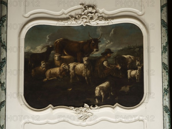 Shepherd's Scene with a Shepherd Milking a Goat. The overall tones are browns and dark blues. The surface is fully composed, the focal point is a cow standing at the back and a group of five animals—three sheep and two goats—filling the right side of the scene. On the right, a seated shepherd in a sheepskin coat and hat milks a goat into a wooden bucket. Next to the goat is a small white kid. At the front a dog laps water from a shallow pond. In the background on the right, city buildings are visible on a hill beneath a dark, puffy-clouded sky. In the upper corner is the Temple of the Sibyl in Tivoli. Several sheep and rams share identical faces. Philipp Peter Roos, known as Rosa di Tivoli born August 30, 1657 in Sankt Goar; died January 17, 1706 in Rome — German landscape painter; the painting may be by him possibly related to NB 4918, NB 4920, and NB 4921. 18th century, Italy; overdoor; oil on canvas; height 91.0 cm, width 127.0 cm., A painted scene set within an ornate white frame s...