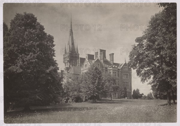 Lentvaris palace; photographer Jan Bulhak 1876–1950; architecture; Lentvaris, Lithuania; palaces; brick architecture; ca. 1934; nature photography/photography; height 11.7 cm, width 16.8 cm; purchase provenance., A large stone building with multiple steep roofs and a tall pointed central spire sits partly obscured by a group of trees in the foreground; several chimneys and decorative roof details are visible on the building. The scene shows a grassy lawn with scattered small flowers in front, bordered by a few large mature trees on both the left and right sides. The sky above is light with subtle cloud texture. Visible colors include shades of gray, black, and white.