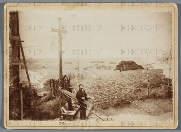 Painting the Raclawice Panorama: Tadeusz Popiel and Ludwig Boller standing with palettes on a scaffolding next to a part of the canvas depicting a cross at the Dziemierzyce crossroads; photographic print, glued to cardboard backing; 1893/1894; Lviv; height 11.7 cm, width 17.4 cm backing: 13.5 x 18.5 cm., The image shows an outdoor scene with a few men standing near wooden structures and a tall wooden post with a crossbar. The men wear dark clothing and hats and stand on or beside a simple wooden platform and ladders. The foreground includes a rough grassy or plowed area with scattered sticks and small trees with thin trunks. In the midground there is a small cluster of low thatch-roofed structures and a few isolated bushes. The background shows a distant, flat horizon with pale sky. Visible colors include sepia tones, brown, beige, cream, and muted gray.