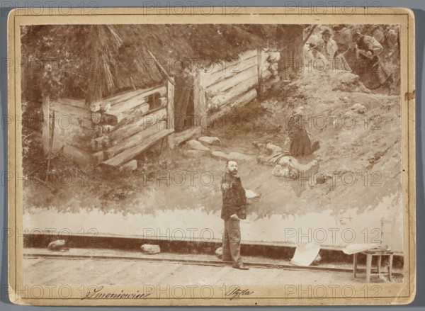 Painting the Raclawice Panorama: Jan Styka standing with a palette next to a part of the canvas depicting a woman mourning a killed peasant, in front of a cottage in Smoniowice at the Dziemierzyce crossroads. Hamel i Feigl Lviv; typesetting of photographic materials; 1892–ca.1905 publisher. Reproduction of painting; Styka, Jan 1858–1925 and others; Kossak, Wojciech 1856–1942 reproduction. Raclawice Panorama, Polish painting; painters' studios; portraits of artists; painting panoramas; painting accessories; painting, 1893/1894; Lviv. Photography from nature; photographic print: height 11.8 cm, width 17.2 cm; print glued onto cardboard backing: backing height 13.5 cm, width 18.5 cm., A sepia-toned scene shows a large painted backdrop of a rustic outdoor setting with rough wooden logs forming a structure and a thatched roof, textured ground with rocks and vegetation, and a few women in period clothing on a slope, one seated and another standing nearby; in front of the painting stands a ma...