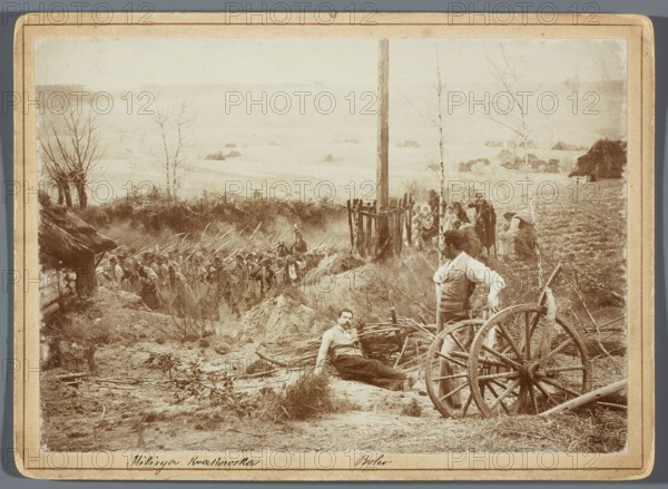 Painting the Raclawice Panorama: Tadeusz Popiel and Ludwig Boller on the artificial terrain next to the part of the canvas depicting the cross at the Dziemierzyce crossroads; photographers Hamel and Feigl; ca. 1892–1905. Subjects: Raclawice Panorama, painters' studios, painting panoramas, portraits of artists. Makers/people depicted: Popiel, Tadeusz 1863–1913; Boller, Ludwig 1862–1896; Kossak, Wojciech 1856–1942 reproduction; Styka, Jan 1858–1925 reproduction. Technique/materials: photograph nature photography, photographic print glued with the entire surface onto a cardboard backing. Date/place: May 1894, Lviv. Dimensions: photographic print h. 11.9 cm × w. 16.9 cm; cardboard h. 13.5 cm × w. 18.5 cm., The image shows a sepia-toned outdoor scene with a group of people and various objects in a rural setting. In the foreground there is a wooden wagon wheel and broken wooden beams beside a standing man wearing light-colored clothing and an apron, and a seated man reclining on the ground w...