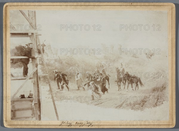 Painting the Raclawice Panorama: Teodor Axentowicz standing on scaffolding by a part of the canvas depicting the escorting of Russian prisoners of war; photographic print, around 1893–1894; print glued to cardboard backing; photography; photographic print height 11.9 cm, width 17 cm; support height 13.5 cm, width 18.5 cm. Artists depicted or associated: Teodor Axentowicz 1859–1938, Zygmunt Rozwadowski 1870–1950, Wojciech Kossak 1856–1942, Jan Styka 1858–1925., A faded sepia-toned scene shows a landscape with a wooden scaffold or ladder structure on the left where a man in dark clothing leans and looks toward the center; a group of people wearing varied garments are walking or standing across the middle ground, some holding long objects and one person appears to be wearing a light-colored cloak; a few mounted riders on dark horses stand to the right of the group; the ground is uneven with patches of grass and a dirt path leading into the distance; the sky and distant background are pale...