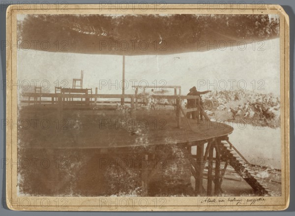 Painting the Raclawice Panorama: painters viewing from a central platform part of a canvas depicting the scene of the capture of a battery of Russian cannons by the leading group of the first column of scythe-bearers, Hamel and Feigl; photographic materials typeset; ca. 1892–1905. Painting reproduction of the Panorama of the Battle of Raclawice by Jan Styka and others; reproduction of Wojciech Kossak. Subjects: artists' studios, painters' studios, painting accessories, painting panoramas, scaffolding, painters. Technique/format: photographic print glued to cardboard backing; photography from nature. Dimensions: photographic print height 11.9 cm, width 17.1 cm; backing height 13.5 cm, width 18.5 cm. Place of origin: Lviv. Date: 1893–1894., The image shows a sepia-toned photograph of a large round wooden platform with railings and a central pole supporting a canopy overhead; on the platform are a few wooden chairs and simple fencing, and a set of wooden stairs leads down from the edge. A...