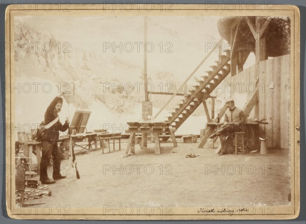 Wojciech Kossak sitting at the central platform of the rotunda and making a sketch for a painting from a model posing in a Russian rifleman’s uniform; Hamel and Feigl, photographic print, 1893/1894, print glued to cardboard print: 12.0 × 17.3 cm; cardboard: 13.5 × 18.5 cm. Depicts painters’ studio with scaffolding and painting accessories during work on the Panorama of the Battle of Raclawice; includes portrait of Kossak, painting models, and references to Jan Styka; photographic documentation of Polish painting and panoramic painting practices artists: Kossak, Wojciech 1856–1942; Styka, Jan 1858–1925., The sepia-toned image shows an outdoor work area with wooden tables and benches, a wooden staircase leading up to a platform on the right, and a tall vertical post near the center. On the left a person wearing dark clothing and a light strap across a shoulder stands holding a long object and a brush or tool above a tripod easel; on the right another person sits on a bench or crate near ...