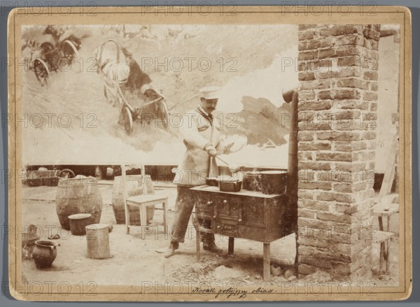 Painting the Raclawice Panorama: Wojciech Kossak with a palette in his hand cooking dinner, in a makeshift kitchen in the rotunda; photographic print, glued to cardboard backing; 1893/1894; photography from nature; print: 11.9 x 17.3 cm, cardboard: 13.5 x 18.5 cm. Depicts Kossak Wojciech, 1856–1942 and painters including Jan Styka 1858–1925 in a painters' studio during work on the Panorama of the Battle of Raclawice; shows painting accessories, palettes, and a kitchen setup. Photographers: Hamel and Feigl Lviv; photographic materials typesetting; active ca. 1892–ca. 1905. Technique: painting reproduction, photography., A sepia-toned scene shows a person wearing a light-colored coat and hat standing by a metal cooking range, holding a utensil over pots and pans on the stove; next to the range is a tall brick chimney. In the foreground are wooden barrels, a simple wooden chair, a few metal containers and a ceramic pot on the ground. Behind the person a painted backdrop with illustrations...