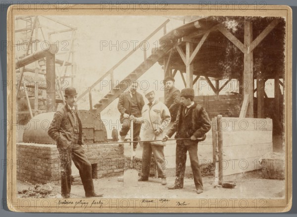 Painting the Raclawice Panorama: painters mixing paints in the rotunda; from the left: Zygmunt Rozwadowski, Ludwig Boller, Wojciech Kossak, Jan Styka, Tadeusz Popiel, Hamel and Feigl, photographic print, ca. 1893–1894; print glued to cardboard backing. Photography of artists in a studio depicting painting panoramas, scaffolding, painting accessories; dimensions of photographic print 11.8 × 17.4 cm, cardboard 13.5 × 18.5 cm., A sepia-toned photograph shows a group of men standing and posing in an outdoor industrial or construction setting with wooden beams and a raised wooden platform with stairs in the background; a few men wear hats including a bowler and a fur hat, and their clothing includes coats and suits in muted tones; one man leans against a low brick structure near a cylindrical metal drum, another man holds a shovel, and another holds a long metal rod across a low wooden barrier; there are wooden posts, planks, and scaffolding visible, with dirt or gravel on the ground, and t...