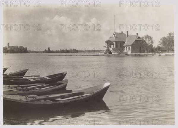 Trakai — shelter; photographer: Jan Bulhak 1876–1950; subject: shelter, lakes, landscapes in Trakai, Lithuania; genre: nature photography; date: ca. 1934; dimensions: 11.4 × 16.2 cm., A lakeshore scene in muted sepia tones shows a group of wooden rowboats clustered in the foreground along the water's edge, their dark hulls and lighter interior seats visible. The calm water reflects soft ripples and the pale sky above. Across the water, a large wooden house with multiple rooflines and a tall flagpole stands on a small stretch of land, with a few trees nearby and a low dock extending into the lake. In the distant background, a low tree line and an indistinct structure sit along the horizon beneath a sky filled with light, fluffy clouds.