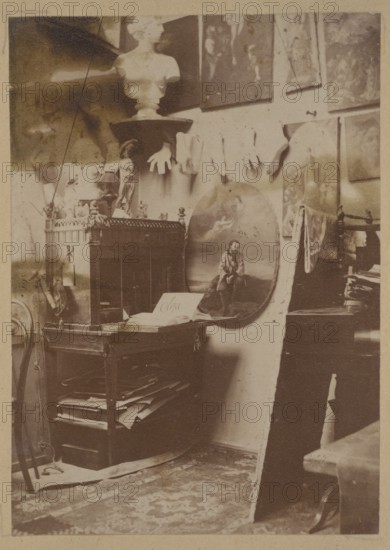 Ignacy Jasinski's 1833–1878 painting studio at the Hotel Europejski in Warsaw; Ignacy Jasinski 1833–1878, photograph, print glued across the entire surface on the verso page of the 22nd page of an album, 1869–1878; subject: interiors, artists' studio, painters; medium: photography nature photography/photograph in an album; dimensions: height 10.3 cm, width 7.3 cm; Polish painting reproduction/iconography., A cluttered interior scene with a wooden table or cabinet holding books and papers, a round painted panel leaning against the wall depicting a seated figure, a bust on a pedestal near the top, several gloves hanging on the wall, framed artworks arranged on the wall, a few sculptural objects and small statuettes on the furniture, a patterned rug on the floor, and visible tones of brown, beige, sepia, cream, and hints of dark gray.