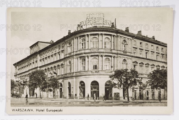 Hotel Europejski at Krakowskie Przedmiescie, Warsaw; photographer, published by Wydawnictwo Salonu Malarzy Polskich Kraków; architecture; hotels; Hotel Europejski Warsaw; Warsaw Masovian Voivodeship; Polish architecture; neon signs; ; postcard glued to the backing with two strips on the shorter side; 1939/1940; Krakow; postcard; height 9 cm; width 14 cm, A black-and-white photograph of a large multi-story corner building with classical architectural features, including arched ground-floor openings, tall rectangular and arched windows, pilasters, and decorative cornices; a rooftop metal sign structure is visible atop the curved corner section. The facade shows a series of repeated window bays extending along both street-facing sides, with a few trees lining the sidewalk in front of the building and a group of people walking or standing near the entrances. The street in front appears wet and reflects light, and the overall scene includes tones of black, white, and various shades of gray.