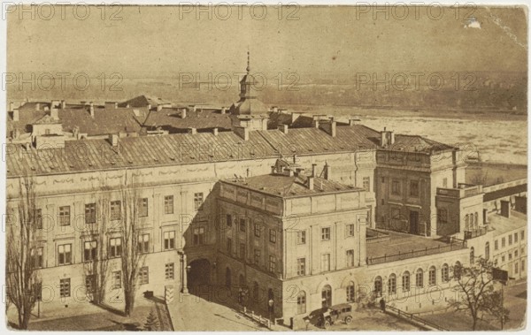 Warsaw. Royal Castle - View from the Congress Side, Jan Bulhak 1876–1950, photographer; printed by National Printing House Krakow, postcard, 1918–1939; depicts Royal Castle Warsaw and Nowy Zjazd street, Warsaw Masovian Voivodeship., A sepia-toned aerial view shows a group of connected classical buildings with pitched and flat roofs, decorative cornices, and rows of windows; a central building with a small tower and spire rises above the surrounding roofs. In the foreground there is a courtyard area with arched ground-floor openings and a few vehicles parked near an entrance, and several tall, leafless trees cast shadows along the façades. The background reveals a distant flat landscape and a shoreline. The visible colors are shades of brown, tan, and beige.