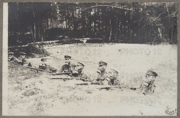 Polish Legions. Soldiers in a trench wearing gas masks Portfolio Polish Legions 1914-1918, part I, unknown author/photographer, World War 1914-1918, trenches, gas masks, wars, Polish Legions 1914-1918, soldiers, photograph glued to cardboard with the entire surface, 1914–ca. 1918, photography, photography from nature, photographic print: height 9 cm, width 13.8 cm, support: height 16.7 cm, width 24.9 cm, A group of people wearing hats and gas masks is lying in a grassy, pale field with rifles held in front of them, arranged roughly in a line; behind them is a darker area of trees and woods with vertical trunks and shaded foliage. The visible colors include shades of black, gray, white, and muted beige.