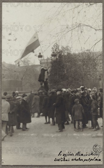 Planting a white-and-red flag in front of the Governor's Palace by a student of the University of Warsaw, after the Germans left the city; photographer: Waclaw Saryusz-Wolski 1870–1933; World War 1914–1918; Warsaw, Governor's Palace, Krakowskie Przedmiescie; Year: 1918; genre scene; national flags Poland; University of Warsaw founded 1816; photograph on postcard paper glued with upper edge to cardboard; board: Polish Legions 1914–1918, part II, 10/11/1918; photographic print: height 13.9 cm, width 8.6 cm; base: height 24.6 cm, width 16.7 cm., A group of people gathered outdoors on a street or square, most wearing dark coats and hats, with a few lighter-colored coats visible; several adults and some children are standing together, some facing different directions. In the background there are leafless tree branches extending across the upper part of the scene and a dense group of trees or shrubs behind the crowd. A tall lamp post is visible with a person standing or climbing on it, and a...