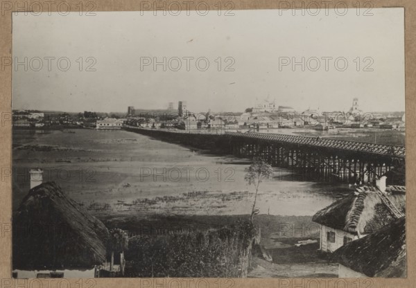 Lutsk — view of the city from the Styr River; photographer, ca. 1918 negative, ca. 1918–1922 print. Photograph photographic print depicting Polish architecture, churches, bridges, brick architecture, river Styr, city views of Lutsk; nature photography. Photographic print: height 10.8 cm, width 16.1 cm; mounted on cardboard: height 35.4 cm, width 25.3 cm. Maker/author: Biske, Ryszard 1895–1938 noted in association with the work., A sepia-toned scene shows a riverside town with a long wooden bridge extending across the water toward a cluster of low buildings and a few larger structures on the far bank. In the foreground there are thatched-roof cottages with dark straw textures and a pale chimney, a narrow dirt path, and a lone slender tree near the water's edge. The river appears shallow in places with visible mudflats and reflective patches, and the bridge is supported by a series of dark wooden piers. The sky is pale and empty above the town, and overall colors visible are shades of br...