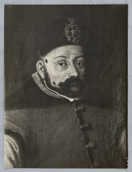 Photograph of a painting: portrait of Stefan Batory, unknown creator/photographer; subject: Stefan Batory King of Poland; 1533–1586 — iconography; male portrait of a ruler; photograph of a work of art; 1st half of the 20th century; dimensions: height 21.0 cm, width 16.0 cm., A portrait of a man shown from the chest up, wearing a dark hat with an ornate decorative element at the front and a dark coat with a high collar and visible round fastenings down the front; his face has a mustache and beard, light skin tones, and visible facial blemishes, with neutral expression and direct gaze; the background is dark and plain, and the overall image appears in shades of black, gray, and white.