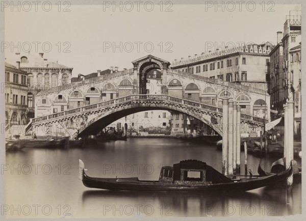 Venice. Grand Canal with a view of the Ponte di Rialto, Carlo Naya 1816–1882, 1865–1875. Photograph photographic print of the Grand Canal and Ponte di Rialto, nature photography; print: 38.5 × 53.5 cm; mounted on cardboard: 50.0 × 67.7 cm., A wide view shows a large stone bridge with arched openings spanning a calm canal, flanked by multi-story buildings with rows of windows and decorative facades; in front of the bridge a long, narrow gondola with an ornate covered cabin floats on smooth water near several tall mooring poles, and a few additional boats and indistinct figures are visible near the bridge and along the quai; the scene is rendered in tones of black, white, and sepia with shades of gray, and the sky above is pale.