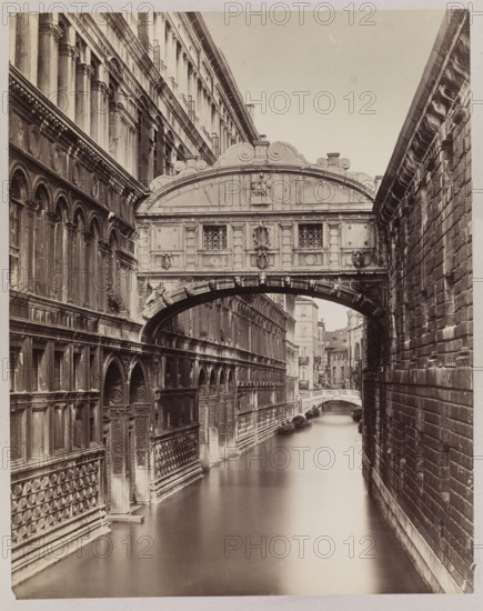 Venice. View of the Ponte dei Sospiri Bridge of Sighs seen from the Ponte della Paglia, Naya, Carlo 1816–1882 photographer/author?, ca. 1870, Venice, photography from nature; photographic print glued to cardboard; photographic print: height 53.5 cm, width 42.0 cm; cardboard: height 68.0 cm, width 49.9 cm., A sepia-toned photograph shows a narrow canal flanked by tall, ornate stone buildings; on the left a building with rows of arched windows, decorative panels, and carved details runs alongside the water, while on the right a textured stone wall rises straight up from the canal. A decorated enclosed bridge with carved reliefs and small latticed windows spans the canal between the buildings, forming a prominent arch over the water. Beyond the arch the canal continues toward a distant view of additional stone buildings and a small arched pedestrian bridge, and a few gondolas or small boats are visible moored along the far edge. The visible colors are shades of brown, beige, and off-white...