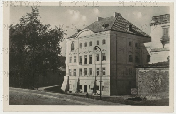 Warsaw. Socialist construction resurrects and discovers the beauty of neglected works of architecture, 1948, photographer Jan Bulhak 1876–1950; postcard; photographic postcard; Palac pod Blacha Palace under the Tin Roof, Warsaw Masovian Voivodeship; height 14 cm, width 9 cm., A street view showing a large multi-story building with a pitched roof and dormer windows next to a smaller section of another building, a tall tree with dense foliage partially obscuring the left side, a curved pathway or road in front, a tall lamppost near the building, a low textured wall or rubble area to the right, a few small figures near the building entrance, and clouds in the sky; visible colors include shades of black, white, gray, and sepia tones.