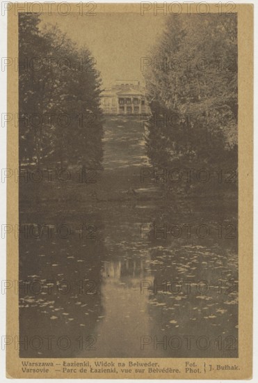 Warsaw. Lazienki, View of the Belweder, ca. 1935. Photographer: Jan Bulhak 1876–1950. Postcard, 14 cm x 9 cm. St. Adalbert's Printing and Bookstore; Polish Society of Railway Bookstores Ruch, Sp. Akc. publisher. Belweder Warsaw., The image shows a distant classical building partially hidden by a group of tall trees on either side, with a long grassy slope leading down to a reflective body of water in the foreground; the scene is rendered in sepia tones of brown, tan, and beige, with darker brown areas for shadows in the trees and lighter tan highlights on the building and water reflections.