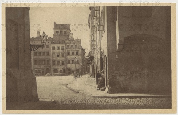 Warsaw. Old Town Market Square, Jan Bulhak 1876–1950 photographer; St. Wojciech's Printing House and Bookstore Poznan printer; Polish Society of Railway Bookstores Ruch publisher; Old Town Market Square, Warsaw; ca. 1930; postcard; 9 × 14 cm, A sepia-toned street scene showing a narrow cobblestone road curving toward a group of multi-story buildings in the distance, with a tall row of buildings lining the right side and a large dark wall or structure on the left foreground; a decorative street lamp projects from the left side near the middle, and several lampposts are visible along the street; a few small human figures stand or sit near the building entrances on the right sidewalk; the image features shades of brown, tan, beige, and dark sepia throughout.
