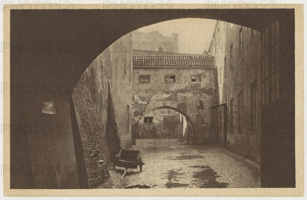 Warsaw. Old Town - Piwna Street, St. Adalbert's Printing and Bookstore; co-creator Bulhak, Jan 1876–1950, photographer; publisher Polish Society of Railway Bookstores Ruch, Warsaw; ca. 1930; postcard; 9 cm x 14 cm, A sepia-toned view of a narrow alley seen through a large archway, with textured walls on either side and a smaller arch further down leading to a walled building with a tiled roof; a simple wooden cart sits near the left wall in the foreground on a rough, patchy ground, and the scene features a range of brown, tan, beige, and darker brown tones throughout.