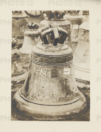 Bell from 1448 from the church in Libusza Galicia requisitioned in 1917/1918 — general view among other bells; bells musical instruments, crafts, looting; war looting; Galicia Austrian partition, Libusza Lesser Poland Voivodeship; war 1914–1918, World War I 1914–1918; Full-surface print re-glued to cardboard, not after 5.10.1918; nature photography, photograph, photographic print: height 10.5 cm, width 7.5 cm; cardboard: height 35.7 cm, width 25.7 cm, A large weathered bell occupies the center of the image with a sculpted crown-like handle on top; behind it are several other bells of varying shapes and sizes partially visible. The bells show textured surfaces with worn areas, decorative bands, and raised relief details. The scene includes a few rounded rims and bases, and a ground surface beneath them. The visible colors are shades of brown, beige, and off-white, with darker brown shadows and lighter beige highlights.