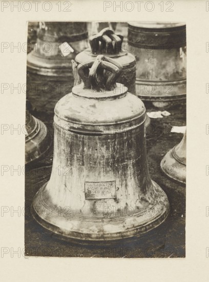 Bell from the church in Siemiechów Galicia from 1557, requisitioned in 1917/1918, unknown creator/photographer, bells, crafts; bell looting during World War I 1914–1918; Galicia Austrian partition. Print glued to cardboard over entire surface; photograph from nature; photographic print: height 10.6 cm, width 7.2 cm; cardboard: height 35.5 cm, width 26.0 cm; not after 5.10.1918., A close-up view of a large weathered metal bell with a rounded body and a decorative crown-like handle on top, positioned in the foreground on a dark surface, with a group of similar bells partially visible in the background; the bell shows surface discoloration, scratches, and patina, and the scene contains shades of brown, beige, cream, black, and gray.