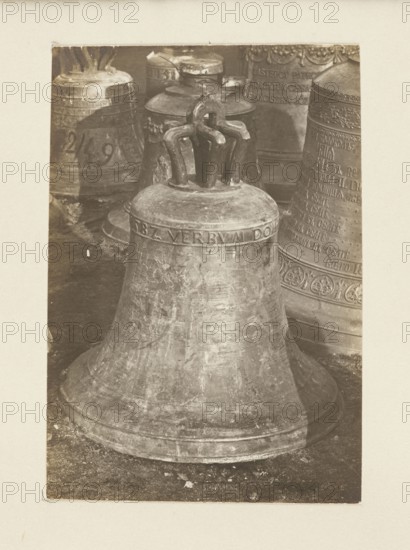Bell from the church in Krynica Galicia, 1582; requisitioned in 1917–1918. Bells; crafts. Plunder during World War I 1914–1918. Krynica Lesser Poland Voivodeship. Photograph from nature; photographic print height 10.6 cm, width 7.8 cm mounted on cardboard height 35.7 cm, width 26.0 cm. Secondary full-surface print glued to cardboard; not after 5 October 1918. Creator/photographer unknown., A large metal bell dominates the foreground with a weathered surface showing scratches and patina in shades of gray and brown; the bell has a rounded flared base and a crown with several curved metal loops at the top. Behind it are a group of other bells of varying sizes and shapes with similar aged textures and decorative bands, visible in tones of gray and muted bronze. The floor beneath the bells appears rough and dark, with scattered small debris in earthy gray and brown tones. The overall image is framed by a light-colored border.