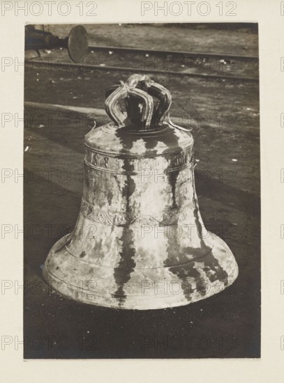 Bell from the church in Glogoczów Galicia, 1583, requisitioned in 1917/1918;, Subject: bells musical instrument; themes: plunder, war World War I. Photograph from nature; photographic print: 10.5 × 7.5 cm; mounted on cardboard: 35.3 × 26.0 cm. Print re-glued on entire surface. Type of place/, The image shows a large metallic bell placed upright on a dark surface, with a decorative crown-like top and ornate banding around its middle; the bell's surface has irregular patches and streaks of lighter and darker tones. Visible colors include shades of black, white, gray, and sepia.