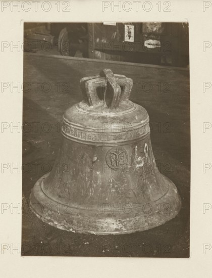 Bell from the church in Lopuszna Galicia from the 11th century, requisitioned in 1917/1918; bells, crafts; war plunder; war 1914–1918, World War I; Lopuszna Lesser Poland Voivodeship; Secondary full-surface print glued to cardboard, not after 5.10.1918; photograph from nature; photographic print: height 10.6 cm, width 7.5 cm; cardboard: height 35.4 cm, width 26.0 cm; /photographer., A large metal bell resting on a flat dark surface fills the foreground, showing a rounded base and a flared skirt with visible wear and scratches. The bell has a sculpted crown with thick looping handles and a raised band around its upper section, and there is a circular emblem on its side. The background shows a darker area with a horizontal ledge and a few indistinct objects behind it. The visible colors are shades of brown, beige, and dark gray.