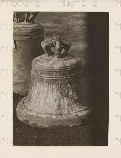 Bell from the church in Osiek near Oswiecim from the 16th century, requisitioned in 1917/1918 – general view; bells, crafts, bells musical instruments; war looting, Galicia Austrian partition; war 1914–1918, World War I; Osiek Lesser Poland Voivodeship, Oswiecim County; Print with the entire surface re-glued onto cardboard, not after 5.10.1918; photograph from nature; photographic print: height 10.5 cm, width 7.5 cm; cardboard: height 35.7 cm, width 26.0 cm, A large metal bell sits on a dark textured ground with a smaller bell partially visible behind it; the bells show worn, weathered surfaces with patina and streaks, and the top of the foreground bell has a sturdy, looped handle with angular supports. The image displays a range of grayscale tones from light to dark, including off-white, beige, gray, brownish-gray, and deep charcoal, with shadowed areas at the right and behind the bells and brighter highlights on the bell rims and tops.