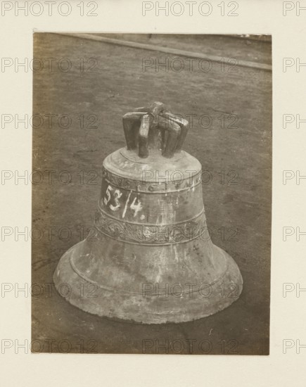 Bell from the church in Powroznik Galicia, 16th century, requisitioned in 1917/1918 — general view; bell; crafts; bells musical instruments; war looting; Galicia Austrian partition; World War I 1914–1918. Print glued to cardboard across entire surface. Photograph from nature; photographic print: height 10.5 cm, width 7.9 cm; cardboard: height 35.3 cm, width 26.0 cm; not after 5.10.1918. Unknown creator/photographer and place of origin., A large metal bell sits upright on a dark, textured ground inside a light-colored rectangular border; the bell has a rounded base, a flared skirt, a decorative band with carved swirls around its middle, and a thick top with a multi-pronged metal fixture. The visible colors are shades of brown, tan, beige, cream, gray, and muted off-white.
