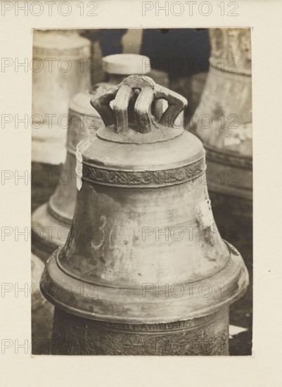 A 16th-century bell from the Gorlice station Galicia requisitioned in 1917/1918 - general view; unknown creator/photographer; bells, crafts, bells musical instruments; plunder, war plunder; Galicia Austrian partition; war 1914–1918, World War I; Gorlice Lesser Poland Voivodeship; Secondary print on the entire surface glued to cardboard, not after 5.10.1918; photograph from nature; photographic print: height 10.5 cm, width 7.2 cm; cardboard: height 35.5 cm, width 26.0 cm, A close-up view of a large metal bell with a textured, worn surface and a decorative band near its top, topped by a thick, sculpted handle; the bell shows patches of lighter and darker tones and a few rough areas, and it sits among a group of other bells and cylindrical objects visible in the background; the image displays shades of brown, beige, tan, cream, and darker brown tones.