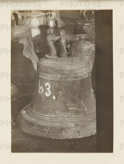 Bell from 1603 from the church in Mogila near Kraków, requisitioned in 1917/1918 - general view; bells; crafts; bells musical instruments; plunder; Galicia Austrian partition; war 1914–1918; war plunder; World War I 1914–1918; Mogila Kraków; Full-surface print re-glued onto cardboard; not after 5.10.1918; photograph from nature; photographic print: height 10.6 cm, width 7.6 cm; cardboard: height 35.7 cm, width 26.0 cm, A large metal bell dominates the foreground with a textured, worn surface and a rounded base, showing a patina of brown and gray tones; its top features a sturdy, sculpted handle with a rope threaded through, and a few lighter scuff marks are visible on the bell's body. Behind it, several other bells and metal objects are partially visible, exhibiting similar aged surfaces and ornate patterns, with colors in muted browns and grays and hints of lighter highlights where the metal catches light. The scene is framed by a dark vertical strip along the right edge and a lighter...