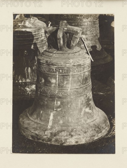 Bell from 1619 from the church in Cerkiew Galicia requisitioned in 1917/1918 — general view; bells; crafts; plunder; war 1914–1918, World War I; Galicia Austrian partition; Print glued on entire-surface cardboard; photograph from nature; photographic print: height 10.6 cm, width 7.9 cm; cardboard: height 35.6 cm, width 26.0 cm; not after 5.10.1918; unknown creator/photographer., A large weathered metal bell dominates the foreground, showing a rounded flared base and a narrower upper section with decorative raised bands and ornamental patterns; the bell's top has a stout, worn handle structure. Behind it are several other similar bells partially visible, stacked and overlapping, with rough textured surfaces. The scene shows a mix of dark and light tones with colors appearing as shades of brown, tan, beige, and muted gray, and areas of lighter wear and patina on the metal. The background is darker, providing contrast with the lighter worn areas on the bells, and the overall surface benea...