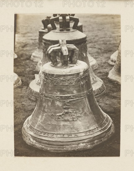 Bell from 1642 from the church in Wieloglowy Galicia requisitioned in 1917/1918 - general view; bells; crafts; bells musical instruments; plunder; Galicia Austrian partition; war plunder; World War I 1914–1918; Print re-glued on the entire surface cardboard; not after 5.10.1918; photograph from nature; photographic print: height 10.5 cm, width 7.9 cm; cardboard: height 35.3 cm, width 25.9 cm, A close-up view of a group of large metal bells arranged in a line on the ground, with the nearest bell in the foreground and others receding into the background; the bells show decorative reliefs and worn surfaces, and each has a heavy crown-like top with integral supporting elements; the ground appears dark and textured; the image tones are sepia and brown with variations of beige and off-white around the bordered edge.