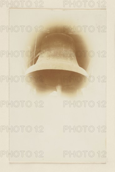 Bell in the parish church in Piotrków – general view, /photographer; bells, crafts; Piotrków Trybunalski Lódz Voivodeship; Print with the entire surface re-glued to cardboard; not after 25/10/1917; photography, photograph of a work of art, photography from nature; photographic print: height 13.9 cm, width 8.8 cm; cardboard: height 31.7 cm, width 23.9 cm., A large bell fills the upper portion of the image, shown from below with its curved lip and interior clapper visible; the bell appears in sepia tones with gradients of brown and beige, and a soft halo of lighter beige surrounds it fading into a pale cream background, with a thin rectangular border framing the scene.