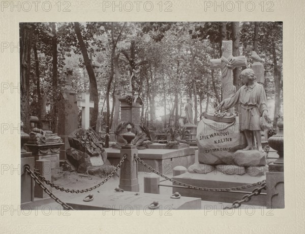 Tombstone of the composer Ignacy Komorowski 1824–1857 at Powazki Cemetery, Warsaw. Photograph glued across the entire surface onto a backing, ca. 1900. Nature photography/photograph, height 17.7 cm, width 24.8 cm. /photographer., The image shows a cemetery scene with a group of stone monuments and statues among trees; a stone sculpture of a young figure stands beside a large carved rock, and a tall cross with draped cloth is visible nearby; several upright tombstones and decorative urns occupy the area, with a low stone wall and chained posts enclosing part of the foreground; the ground is uneven with scattered stones and carved reliefs, and tree trunks and leafy branches form a wooded backdrop. The visible colors are shades of gray, sepia tones, off-white, and muted brown.