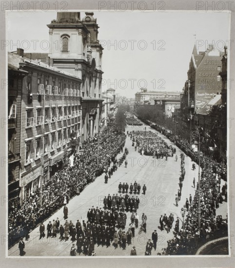 Manifestation on Krakowskie Przedmiescie on the 125th anniversary of the adoption of the Constitution of May 3rd, 03.05.1916. Photographer: Waclaw Saryusz-Wolski 1870–1933. City view/parade/national celebration on Krakowskie Przedmiescie, Warsaw; shows churches St. Krzyza and Hotel Bristol. Photograph from nature, photographic print glued over entire surface onto white paper backing and cardboard no. 6. Date: 1916 World War I. Print: 24 cm × 21.7 cm; backing: 28 cm × 37.5 cm., A black-and-white street scene showing a wide avenue flanked by multi-story buildings with many windows and architectural details, a large church or ornate building on the left with a tower, and a mix of storefronts and taller structures on the right; the avenue is filled with a large procession of people walking in organized groups down the center and dense crowds lining both sidewalks, with some people leaning from windows and balconies; there are a few lampposts along the right sidewalk, several leafless trees...