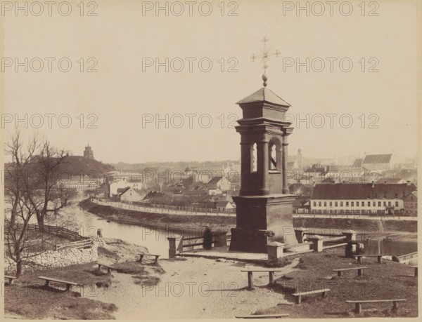 Vilnius - a chapel on Snipiszki, in the background a panorama of the city, Czechowicz, Józef 1818-1888 author, Vilnius Lithuania - photography, Castle Hill Vilnius, panoramas, Snipiszki Vilnius, chapels, View of the city from the Snipiszki suburb, in the foreground a chapel with the miraculous figure of the Crucified Christ from 1720, on the left the arsenal, Castle Hill, the Tyszkiewicz Palace building, in the background the Cathedral, in the background the bell tower of St. Jana., ca. 1875, Vilnius, photograph, photographic print: height 21.0 cm, width 27.5 cm, cardboard: height 27.8 cm, width 34.5 cm, A sepia-toned scene showing a riverside town from an elevated grassy vantage with a stone pedestal structure topped by a decorative metal cross in the foreground; the pedestal has columns and open arches and sits on a paved platform with a low stone railing. Around the foreground are a few simple wooden benches on patches of grass and dirt, and a worn path leading toward the structure....