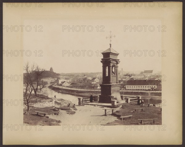 Vilnius - a chapel on Snipiszki, in the background a panorama of the city; Józef Czechowicz 1818–1888 photographer; Vilnius Lithuania - photography; Castle Hill Vilnius; panoramas; Snipiszki Vilnius; chapels; View of the city from the Snipiszki suburb: in the foreground a chapel with the miraculous figure of the Crucified Christ from 1720; on the left the arsenal, Castle Hill, the Tyszkiewicz Palace building; in the background the Cathedral and the bell tower of St. Jana; ca. 1875; photograph, photographic print: 21.0 × 27.5 cm; mounted on cardboard: 27.8 × 34.5 cm., A raised stone monument with an ornate cross at its top stands on a grassy terrace overlooking a river and a town; the monument is dark grey and has an arched niche with a bell visible, surrounded by stone railings and short posts. In the foreground there is a dirt path and a few simple wooden benches arranged on the grassy slope, with patches of bare earth. The river runs left to right behind the monument, bordered by a l...
