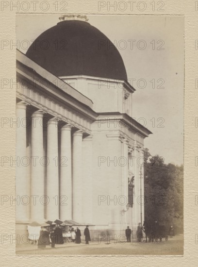 Vilnius. St. Casimir's Chapel; photograph by Jan Bulhak 1876–1950, pictorialism, 1912. Photograph, photographic print: 15.6 × 11.0 cm another print: 15.5 × 11.0 cm; backing: 31 × 22.5 cm; album page: 31.1 × 23.5 cm., A large classical building facade with a dark domed roof and light colored columns creating a colonnade, shown from an angled side view. Along the base of the building several people are standing near a group of market stalls with canopies, and a few individuals are walking or standing nearby. To the right of the building there is a group of trees and a horse-drawn carriage partially visible. The scene includes colors of black, white, cream, beige, brown, and muted green.