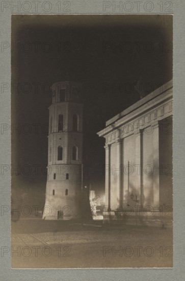 Vilnius. Cathedral at night; photograph by Jan Bulhak 1876–1950, pictorialism, nocturne photography of bell towers and cathedral Vilnius, 1912–1913 print 1914. Photographic print: 16.0 × 10.2 cm printed twice, backing: 31 × 22.5 cm, album page: 31.1 × 23.5 cm., The image shows a night scene with a tall cylindrical tower featuring a few arched openings standing next to a large classical building with columns and an entablature; the tower and building are illuminated against a dark sky, and there is a paved area in front. Visible colors include shades of black, dark gray, light gray, and muted beige tones.
