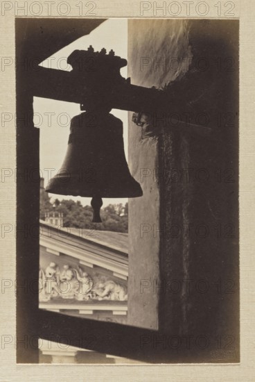 Vilnius. Cathedral bell tower; photograph in the album Vilnius Temples, volume 1, Jan Bulhak 1876–1950, photographer; pictorialism; cathedral Vilnius; photography of bells; 1912; Vilnius; photograph, photographic print: 15.3 × 10.0 cm and 15.5 × 10.0 cm; backing: 31 × 22.5 cm; album page: 31.1 × 23.5 cm, A sepia-toned photograph showing the dark silhouette of a bell hanging from a horizontal beam inside a window-like opening, with the bell's clapper visible and the bell's outline contrasted against a lighter background; to the right a textured vertical wall in shadow runs through the frame, and beyond the opening a portion of an ornate building facade with sculpted relief and a roofline is visible along with a group of trees, all depicted in shades of brown, beige, and dark sepia.
