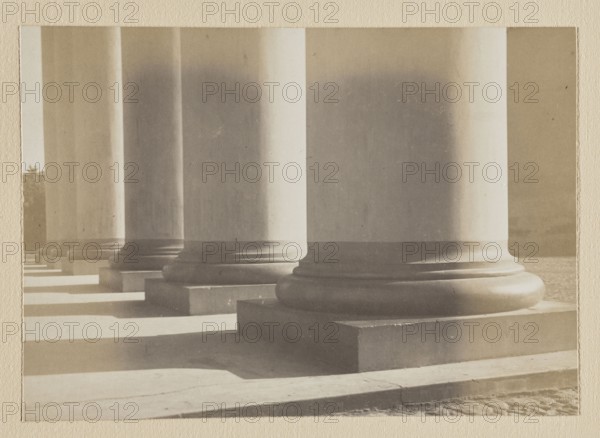 Vilnius. Colonnade of the Cathedral's pediment; photograph in the album Vilnius Temples, volume 1, Bulhak, Jan 1876–1950, pictorialism, Vilnius, photography, cathedral Vilnius, 1912, photograph, photographic print: height 11.5 cm, width 16.4 cm; photographic print: height 11.4 cm, width 16.3 cm; backing: height 30.5 cm, width 22.5 cm; album page: height 31.1 cm, width 23.5 cm, A row of large classical columns seen from a low angle, showing their broad round bases resting on square plinths and casting long rectangular shadows across a stone floor; the columns are light beige to gray with subtle darker shading, the bases are slightly darker gray, and the floor tones range from pale beige to muted gray; the image has a soft, subdued sepia-like overall tint and a textured border around it.
