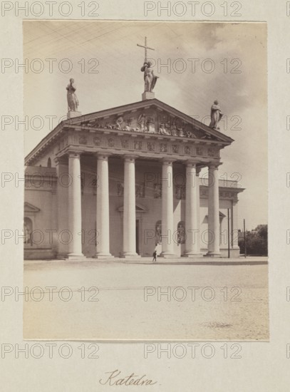 Cathedral; photograph, Bulhak, Jan 1876–1950, Vilnius Lithuania, cathedral Vilnius, 1912, photograph, photographic print: height 14.8 cm, width 11.2 cm, album page: height 31.0 cm, width 23.5 cm, A large classical building with a portico supported by a group of tall columns stands centered in the image, topped by a triangular pediment with sculptural relief and three statues along the roofline, one holding a cross; the façade and columns appear in shades of off-white and light beige, with shadows creating darker beige and gray tones. The sky behind the building shows muted gray and pale beige clouds. In front of the building is a wide open paved area in pale beige, and a small dark figure is visible near the base of the columns. Along the left and right edges of the building are architectural extensions in similar off-white and beige tones, and a few darker vertical elements and foliage appear at the far right in muted green-gray. The overall image has a sepia-like tint with warm beige...