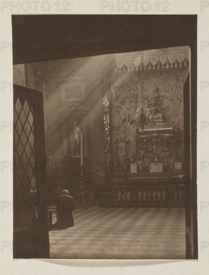 Cathedral. St. Casimir's Chapel; photograph, 1914. Photographer: Jan Bulhak 1876–1950. Subject: St. Casimir's Chapel in the Vilnius Cathedral Baroque architecture. Medium and format: photographic print, height 15.0 cm, width 11.4 cm; mounted on album page, height 31.0 cm, width 23.5 cm., A sepia-toned interior scene shows a person kneeling on a patterned floor in front of an ornate altar area. Sunlight streams through high windows creating visible beams that illuminate dust in the air. The altar wall features sculpted reliefs, a chandelier, decorative columns, and a raised platform with framed objects. A tall candle stand and a statue in a niche are visible near the altar. A partially open doorway with a diamond-patterned glass panel frames the left side of the scene. Colors visible are brown, beige, and muted gold tones.