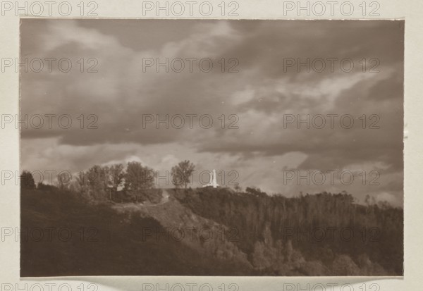 The Three Crosses Mountain with Three Crosses; photograph, Bulhak, Jan 1876–1950, Wiwulski, Antoni 1877–1919, monuments, The Three Crosses Mountain Vilnius, after 1916, Vilnius place of origin, photographic print: height 11.0 cm, width 16.5 cm, album page: height 31.0 cm, width 23.5 cm, A sepia-toned landscape photograph showing a ridge with trees along its crest and a taller cluster of trees near a bright vertical structure on the ridge; the foreground contains a darker wooded slope leading up to the ridge, and the sky above is filled with large, dramatic clouds.